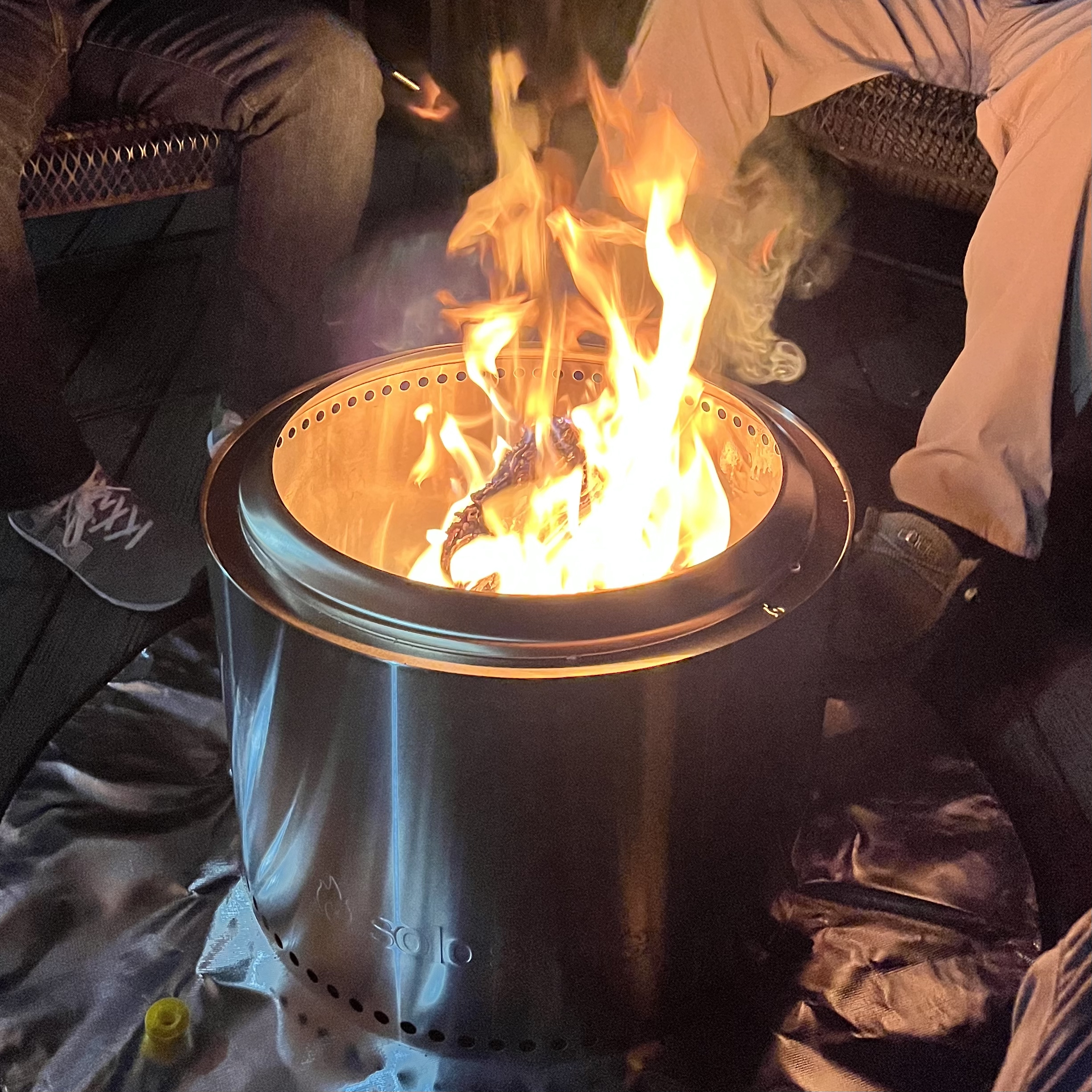 Legs of two men around the brushed stainless steel of the Solo Stove Bonfire with a flames coming up out of the top of the cylinder.